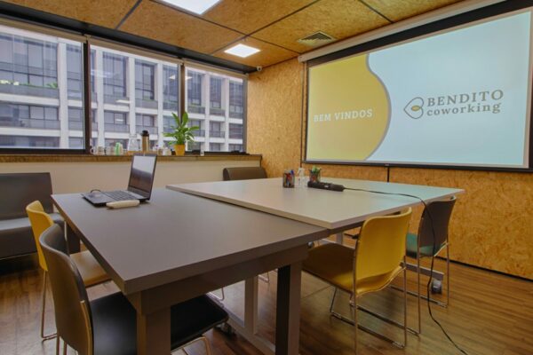 Sala de reunião com uma projeção de bem-vindos ao lado da logo do Bendito Coworking e mesas juntas com 6 lugares