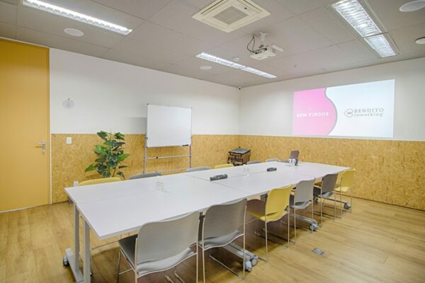 Sala de reunião ampla em coworking, com 1112 lugares, projetor e mesa de fundo, acompanhada por uma lousa branca